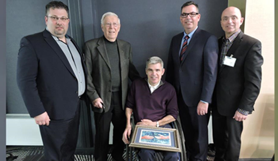 four men standing. one men holding a picture frame is in a wheelchair