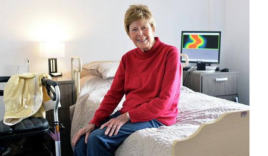 A senior woman is sitting on a bed in front of a computer screen