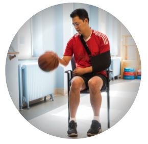 Male patient in red gym wear dribbling a basketball in physio gym with right arm while left arm rests in a sling