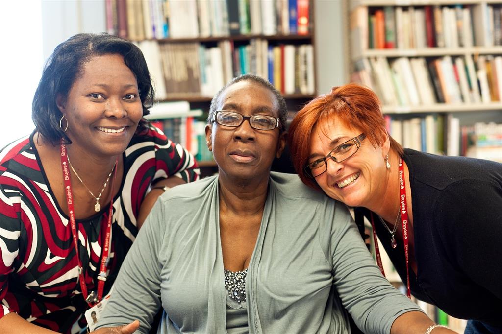 Two female Bruyère employees are couched down to a woman patient in wheelchair smiling a library